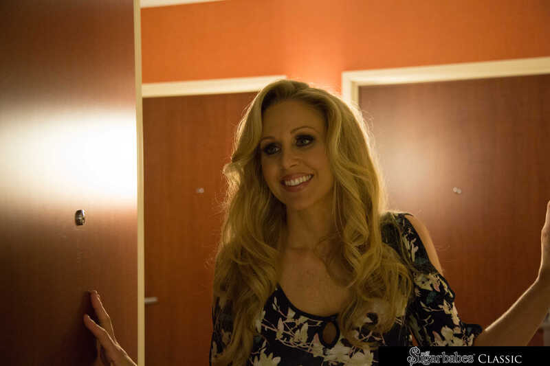 In the Julia Ann Naughty America porn scene 'Julia Ann strips and fucks her sugar daddy before dinner' from Milf Sugar Babes, Julia Ann is standing in a hotel room with a door behind her.