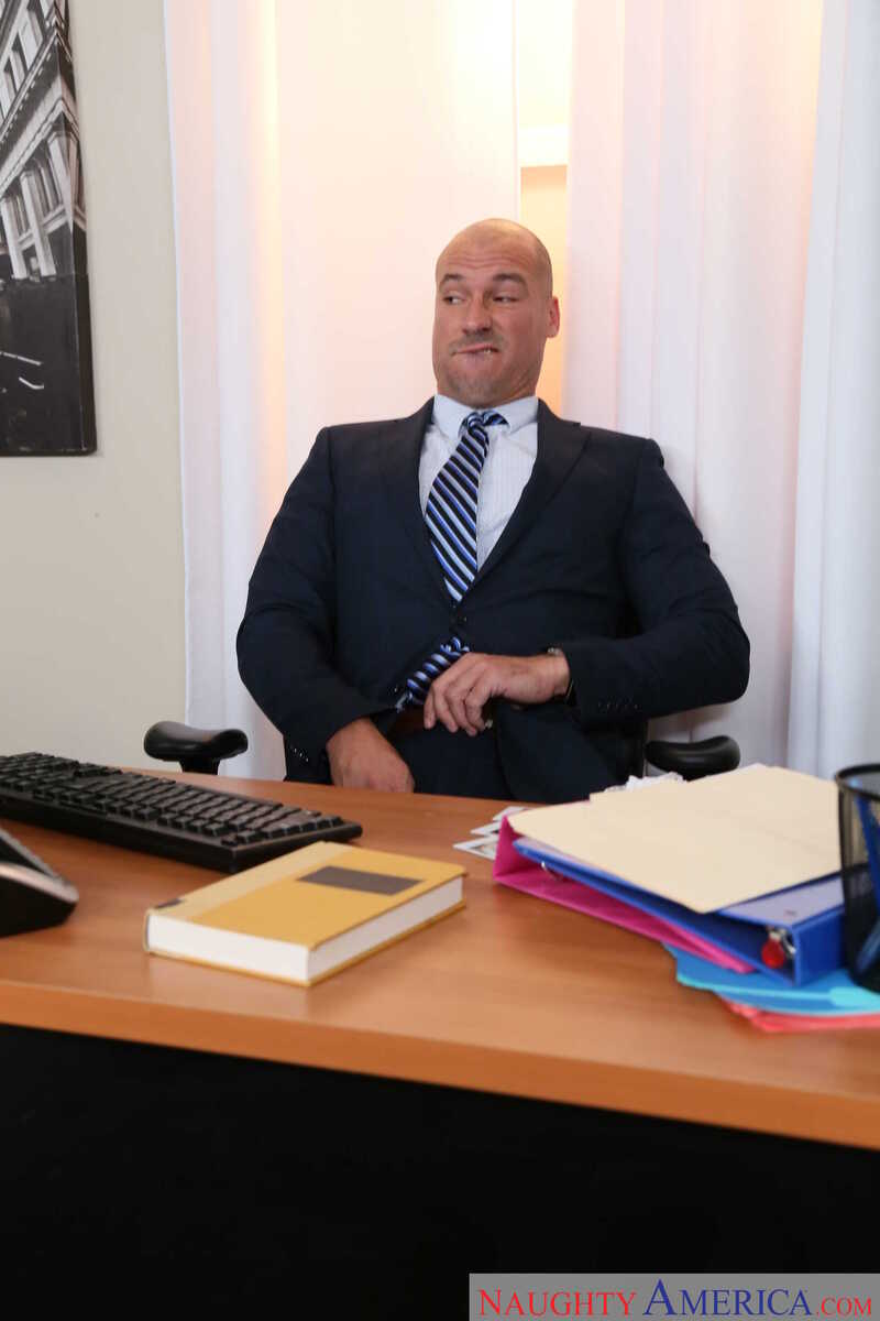 Scene from Naughty America Alex Blake porn video 'Alex Blake Gets Fucked In The Office' where Alex Blake is a bald man in a suit and tie sitting at a desk.