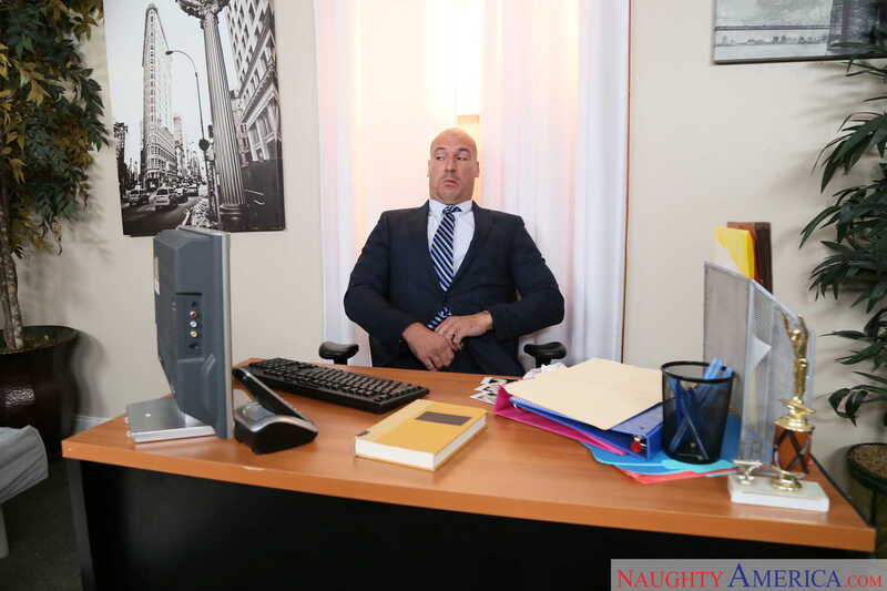 Scene from Naughty America Alex Blake porn video 'Alex Blake Gets Fucked In The Office' where Alex Blake is sitting at his desk in his office.